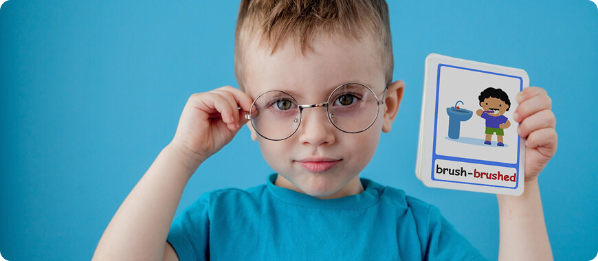 A young boy wearing glasses holds up a flashcard with the word 'brush-brushed, while learning past simple