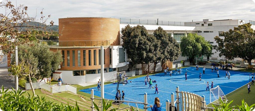 Actividades activas al aire libre en The British School of Barcelona Alumnos practicando deporte en un vibrante campo azul rodeado de árboles y modernos edificios